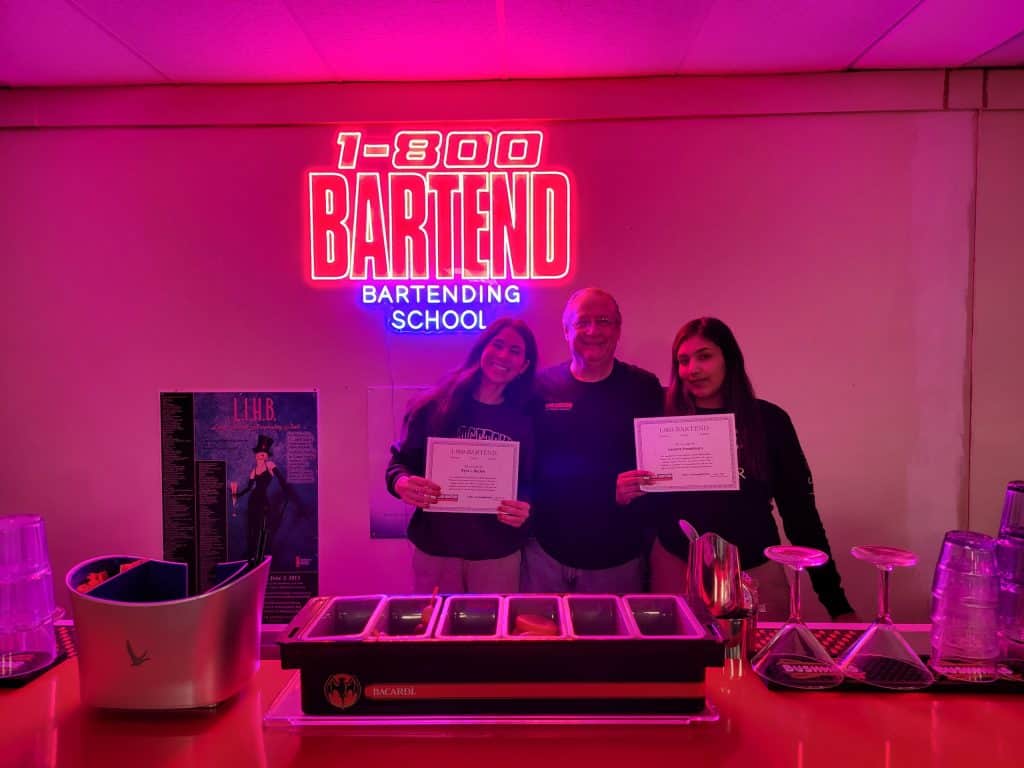 Three people stand behind a bar setup, smiling and holding certificates. A neon sign above them reads "1-800-BARTEND Bartending School." The lighting is pink, with bar tools and glasses in front—perfect graduates from the dynamic New York Bartending School experience.