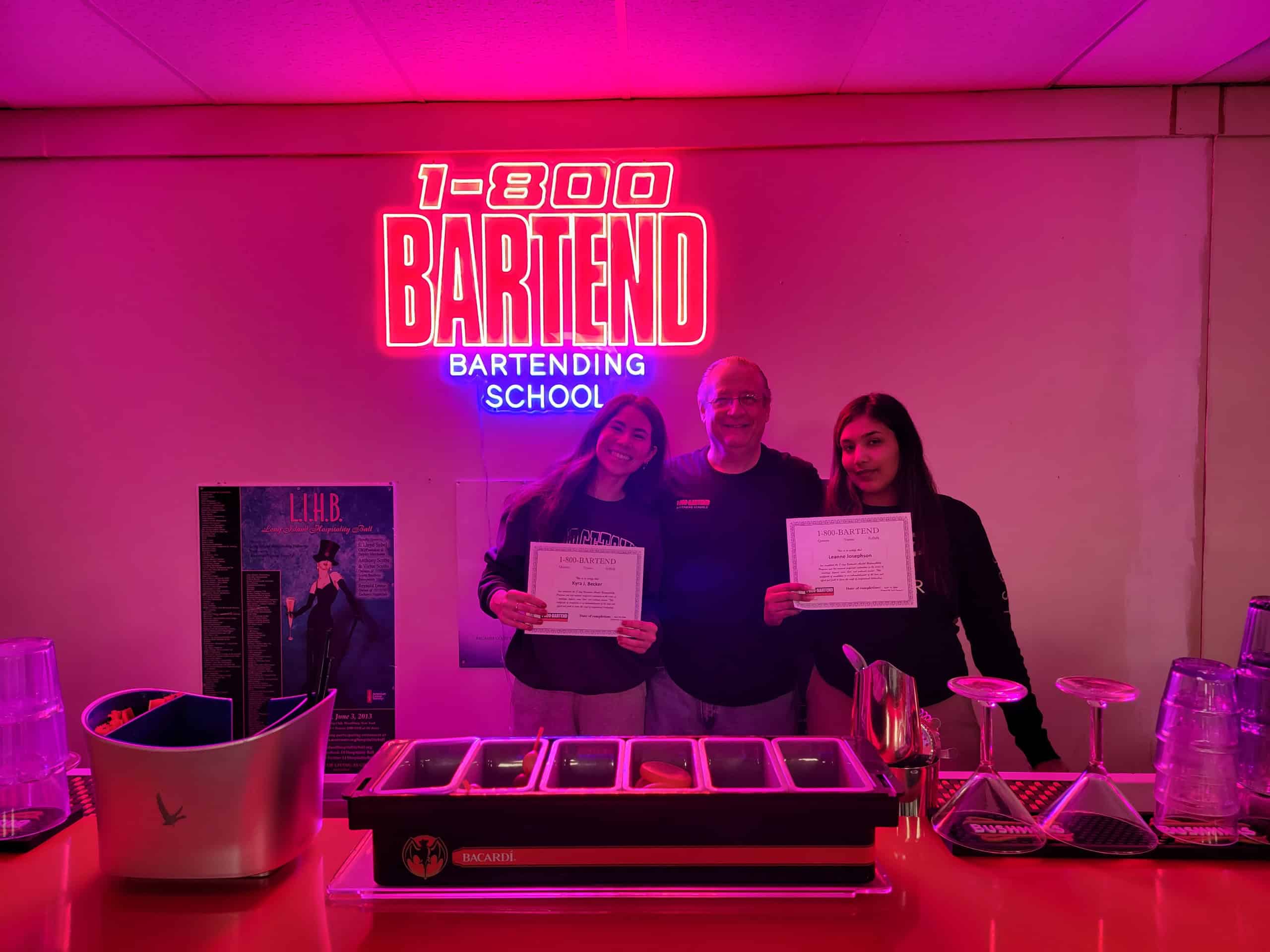 Three people stand behind a bar setup, smiling and holding certificates. A neon sign above them reads "1-800-BARTEND Bartending School." The lighting is pink, with bar tools and glasses in front—perfect graduates from the dynamic New York Bartending School experience.
