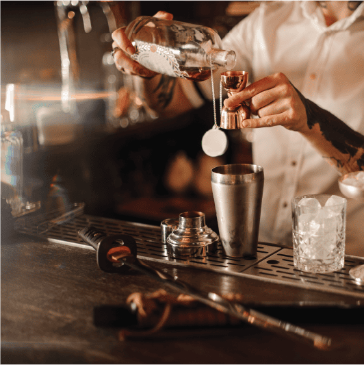 A bartender, trained at Queens Bartending School, pours liquor from a bottle into a jigger over the bar counter. A shaker, glass with ice, and bartending tools adorn the warmly lit setup, creating a cozy atmosphere.