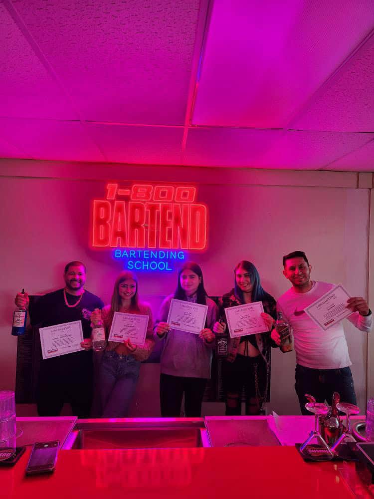 A group of five people posing with certificates and bottles in front of a neon sign that reads "1-800 BARTEND Bartending School." The bar counter is illuminated with pink and purple lighting, capturing the vibrant spirit of the Queens Bartending School.