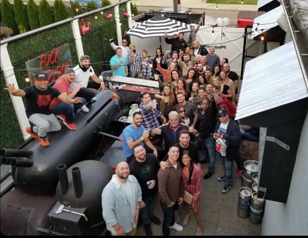 A large group gathers on an outdoor patio, posing for a photo amidst greenery, decorative lights, and a barbecue smoker. Enjoying a casual social event, some have just finished Long Island Bartending School classes, adding flair to the lively atmosphere.