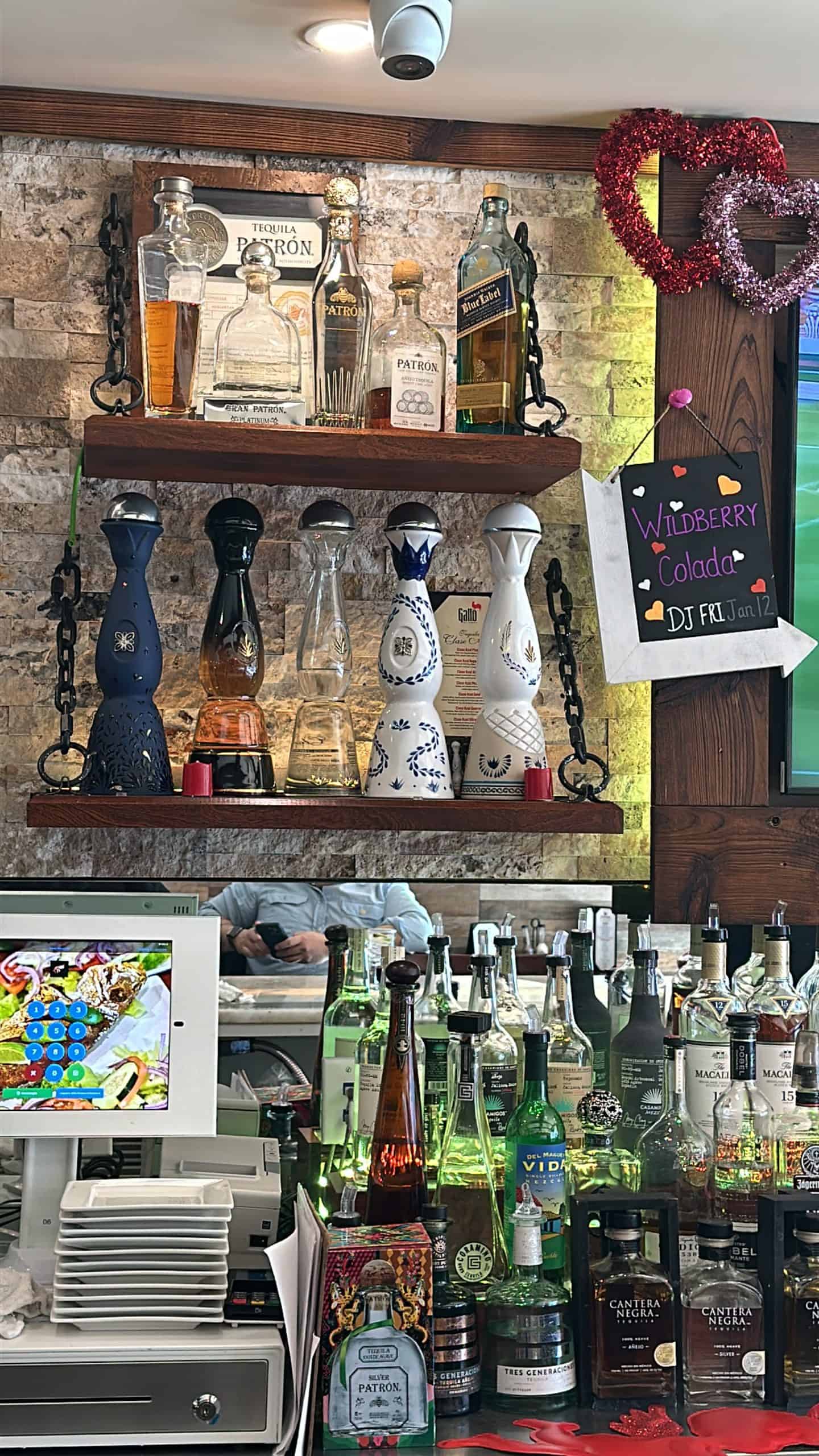 Shelves filled with various tequila bottles are displayed in a bar setting, reminiscent of Long Island Bartending School. A chalkboard sign on the right reads "Wildberry Colada Fri 11am" next to two red heart decorations. Below, a counter with more tequila options is visible, perfect for aspiring mixologists.