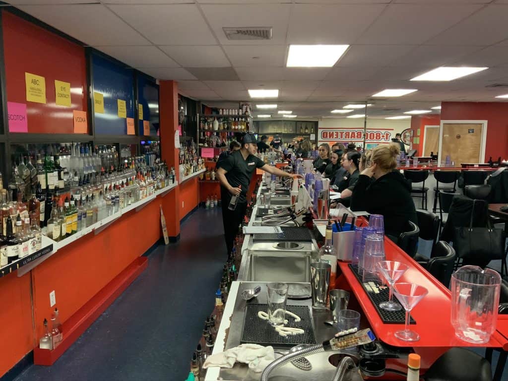 A bartender, perhaps a graduate of Queens Bartending Classes, is crafting a drink behind a long bar counter lined with various bottles. Patrons on stools watch intently in the brightly lit and organized space, featuring a large mirror and red and orange walls.