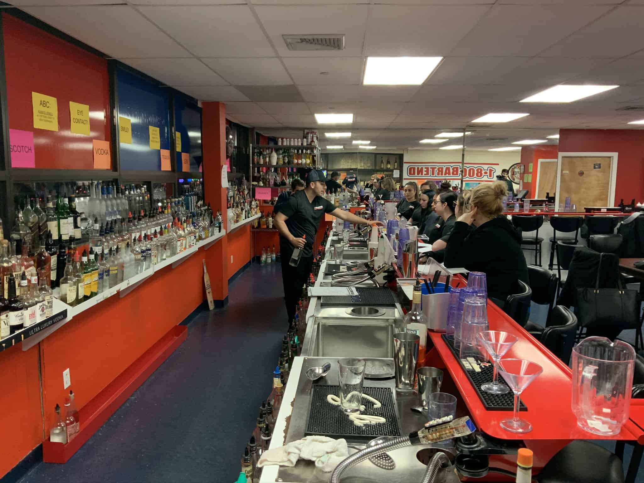 A bartender, perhaps a graduate of Queens Bartending Classes, is crafting a drink behind a long bar counter lined with various bottles. Patrons on stools watch intently in the brightly lit and organized space, featuring a large mirror and red and orange walls.