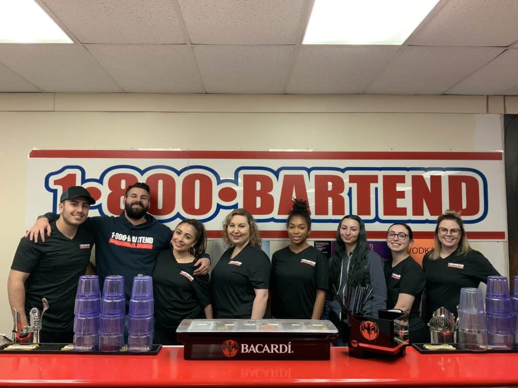A group of eight people wearing matching black shirts stands behind a red bar counter with Bacardi branding, representing the Long Island Bartending School. Behind them is a large sign that reads "1-800-BARTEND." They are smiling at the camera.