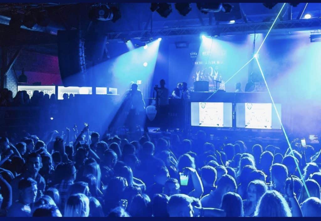 A crowded nightclub with people dancing under vibrant blue lighting and graduates from Queens Bartending School expertly crafting cocktails. A DJ performs on stage with illuminated screens, while laser beams cross the room, enhancing the energetic atmosphere.