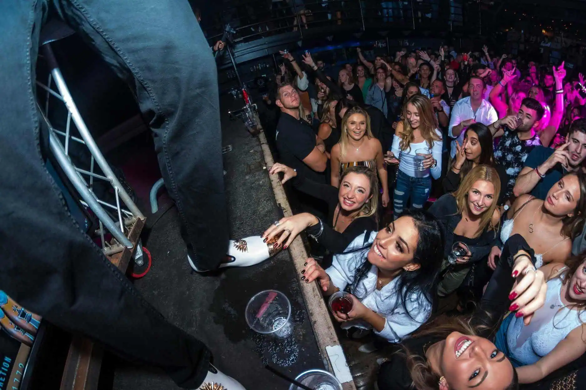 A lively nightclub scene with a crowd of people enjoying a performance unfolds, as someone with skills likely honed at New York Bartending School stands on the bar, interacting with the audience. Drinks and hands are raised, creating an atmosphere that is both vibrant and energetic.