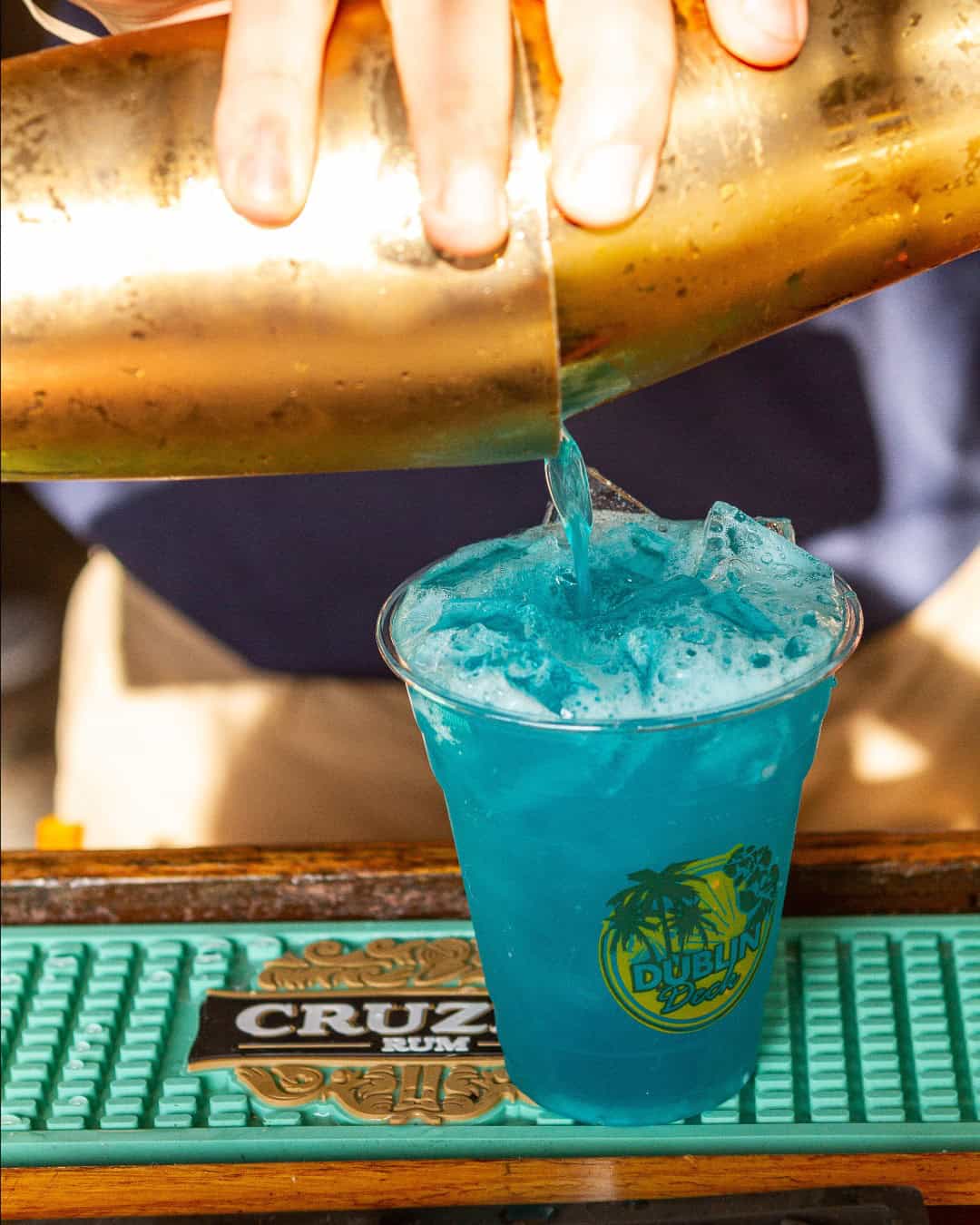 A bartender, fresh from New York Bartending Classes, pours a vibrant blue cocktail from a gold shaker into a plastic cup with ice. The cup features a tropical palm tree logo, set against a green rubber mat displaying "CRUZAN.