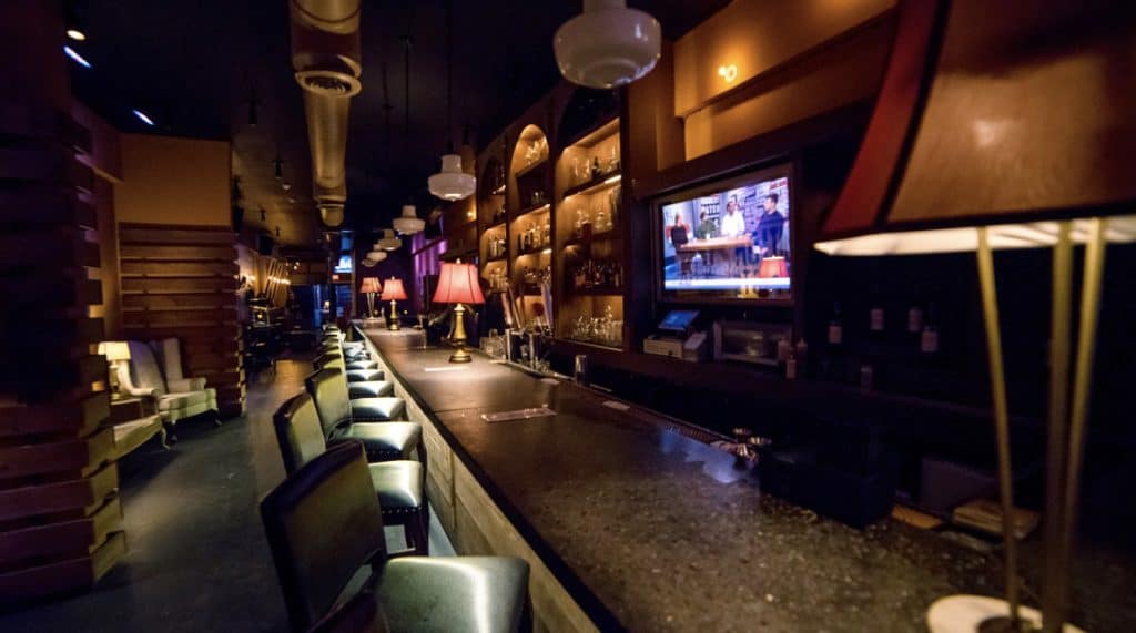 A dimly lit bar interior with a long counter and bar stools captures the essence of New York Bartending Classes. Shelves with bottles and glasses line the wall. A warm, ambient glow emitted by table lamps and overhead lights complements a television screen displaying a show or news.