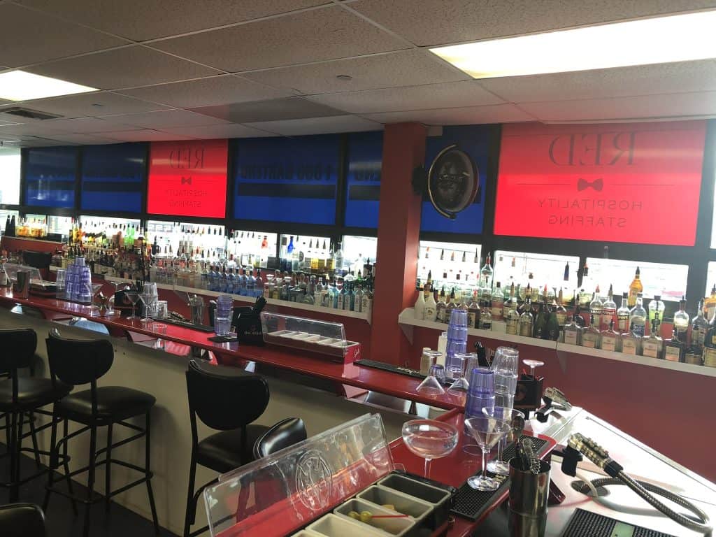 A bar with red and blue signage, featuring a long counter with stools, various bottles of alcohol on shelves, and bar equipment such as glasses and shakers. Perfect for practice if you're taking Queens Bartending Classes. The ceiling is lined with fluorescent lights.