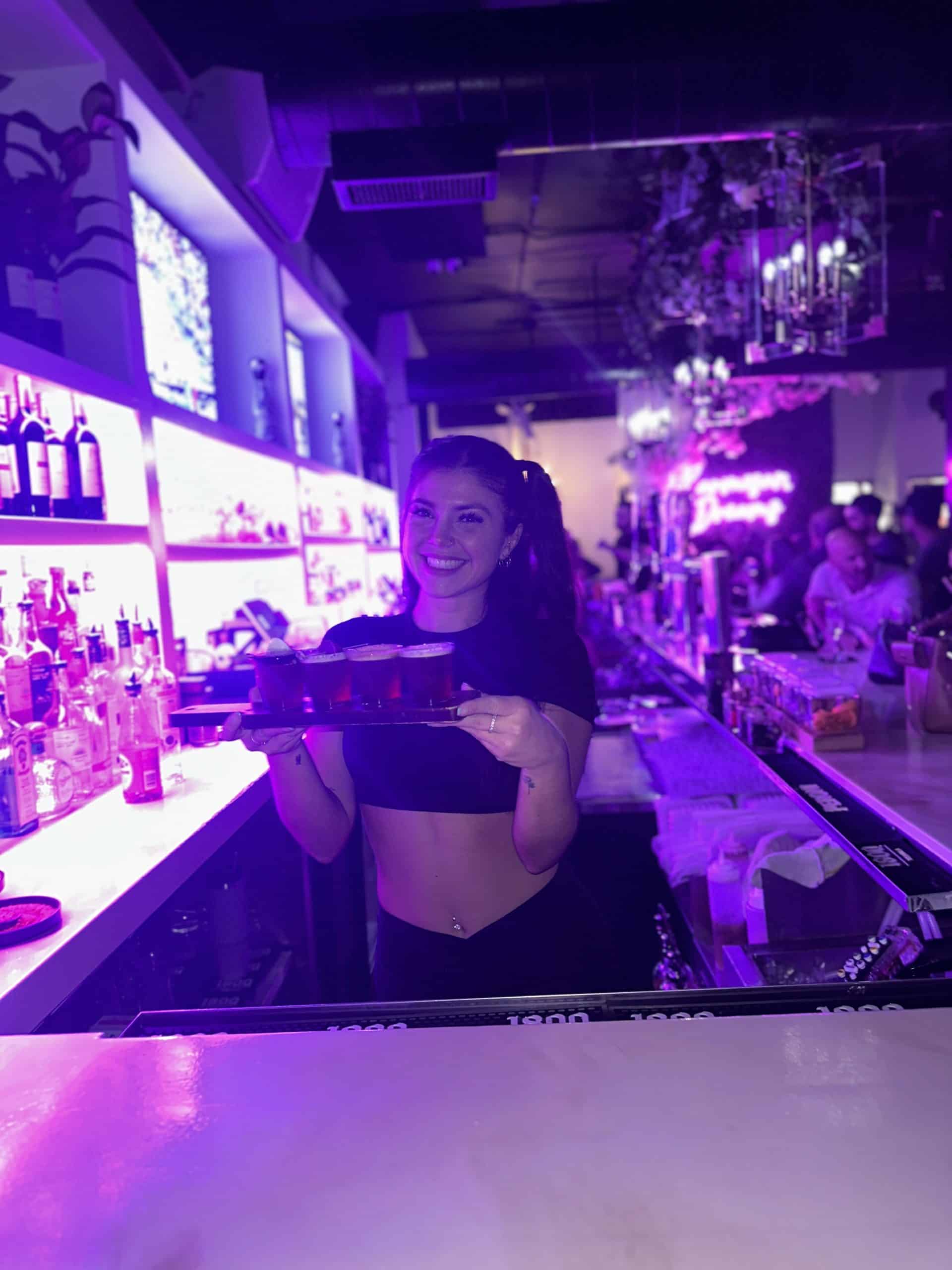 A bartender in a cropped top smiles and holds a tray with glasses at a dimly lit bar with purple lighting. Bottles line the shelves behind her, reflecting skills honed at New York Bartending School, as people in the background create a lively atmosphere.