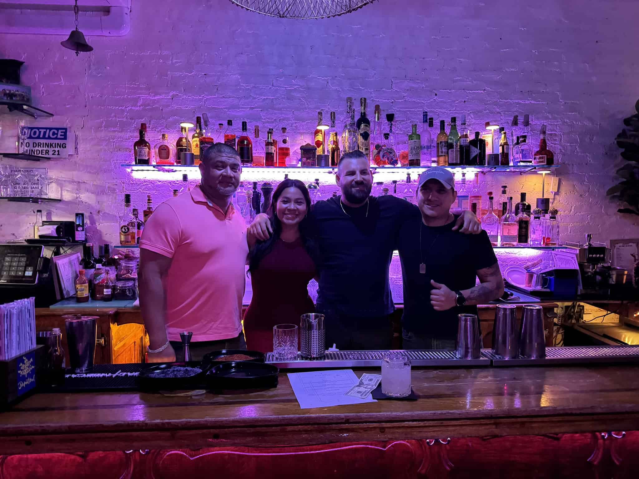 Four people stand smiling behind a bar with bottles and soft purple lighting, embodying the relaxed atmosphere of New York Bartending School. The group—three men and one woman, all casually dressed—reflects the friendly vibe fostered in these popular New York bartending classes.