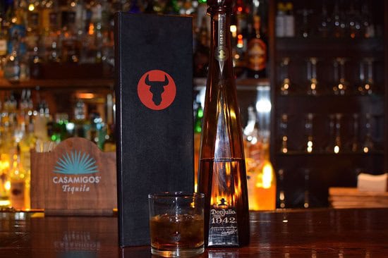 A bottle of Don Julio 1942 tequila sits elegantly on a wooden bar with a filled glass, capturing the essence taught at Long Island Bartending School. Nearby, a sleek black box with a red bull logo completes the scene against blurred shelves of various liquors.