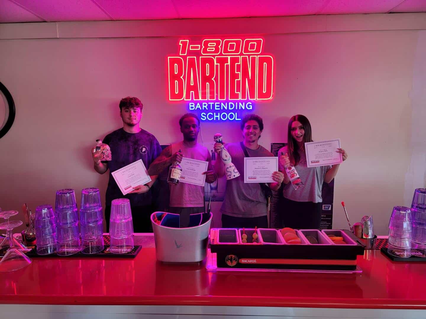 Four people stand behind a bar counter holding certificates and bottles, grinning widely. Above them, a neon sign reads "1-800 BARTEND BARTENDING SCHOOL." Stacked glasses and bar tools pepper the counter, highlighting their recent achievement in Queens Bartending Classes.