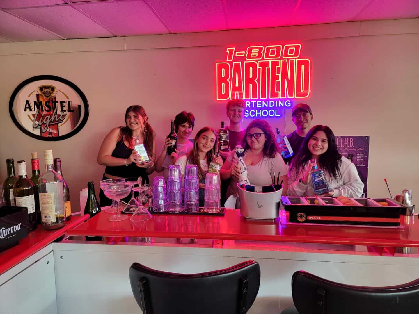A group of people stands behind a bar counter with various bottles and glasses, posing for a photo. A neon sign reads "1-800 BARTEND Bartending School" above them, capturing the vibrant spirit of the Queens Bartending School in training mode.
