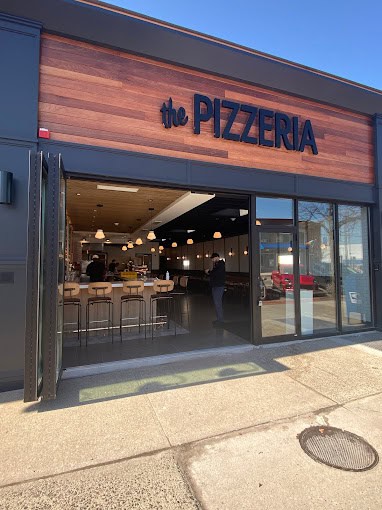 The image shows the entrance to a restaurant called "The Pizzeria." The exterior features a modern design with wood accents. Through the open doors, bar stools and hanging lights are visible, along with a person standing inside.