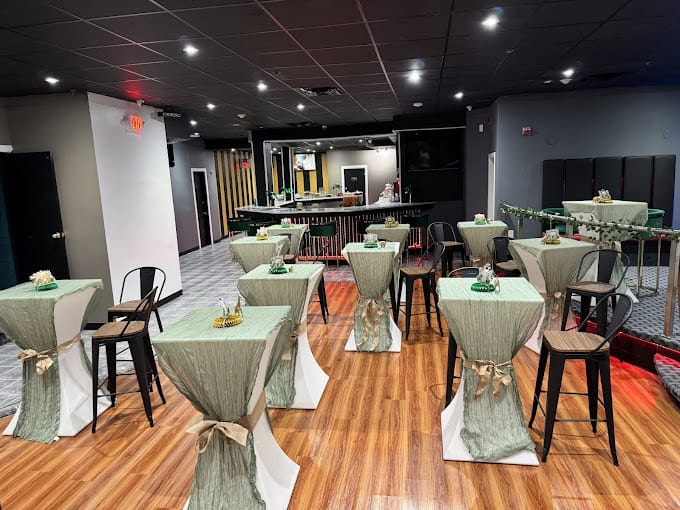 A chic event space with high tables covered in green tablecloths tied with bows. Black chairs surround the tables. The room has a wooden floor and a dark ceiling with spotlights. There's a bar at the back and decorative plants on the tables.