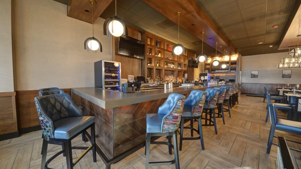 A modern, stylish bar with high-backed chairs and pendant lighting. It features a counter with a decorative pattern, a display of bottles, and a mounted TV. The seating area has wooden accents and contemporary dining tables in the background.