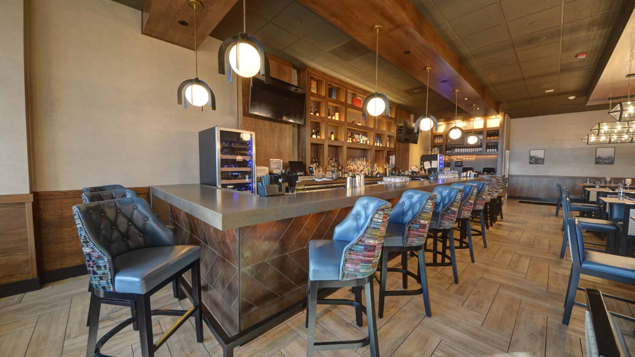 A modern, stylish bar with high-backed chairs and pendant lighting. It features a counter with a decorative pattern, a display of bottles, and a mounted TV. The seating area has wooden accents and contemporary dining tables in the background.