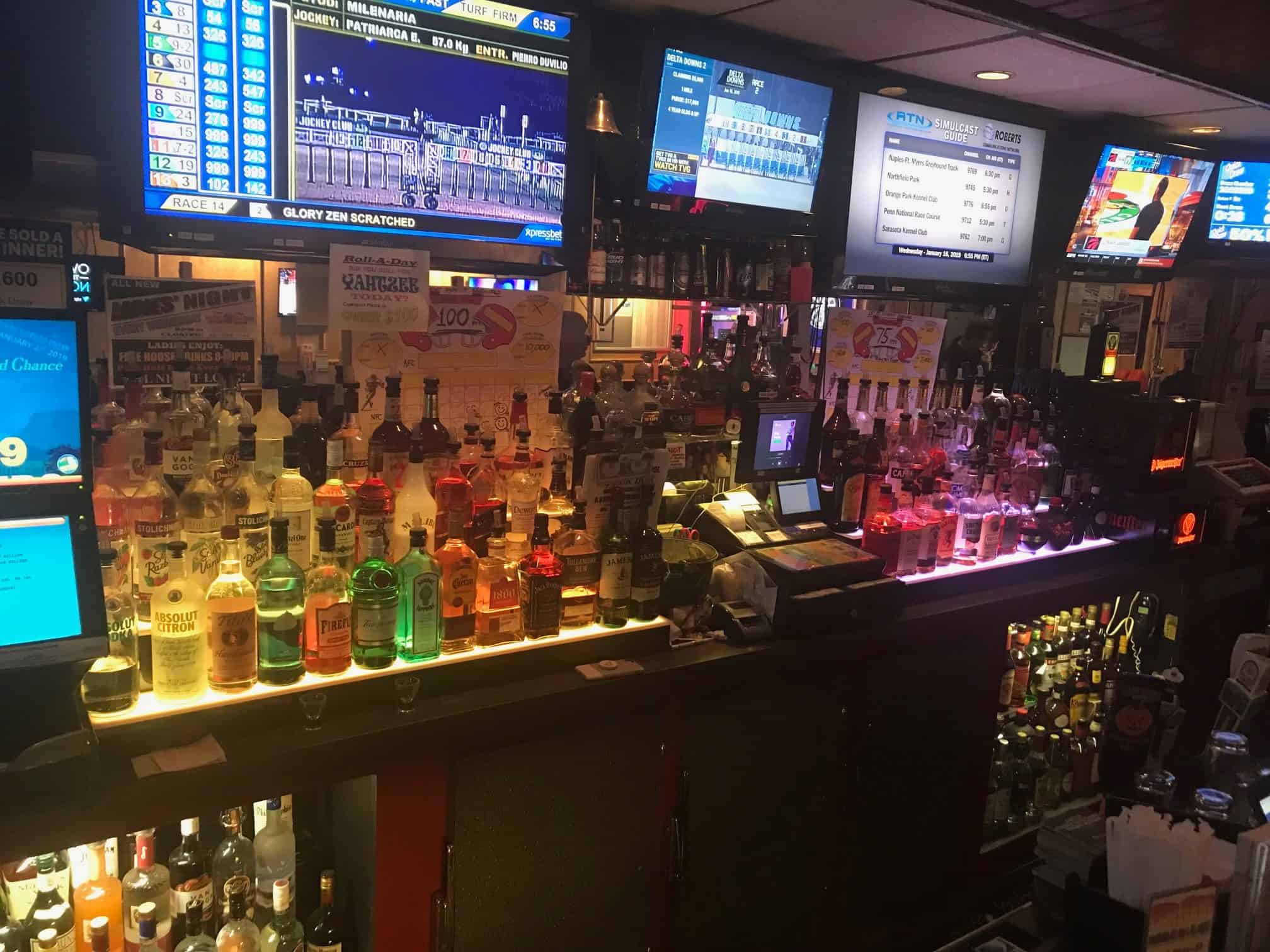 A dimly lit bar counter is lined with various liquor bottles. Several televisions above display sports channels and betting odds. A cash register sits on the counter, surrounded by promotional posters and drink menus.