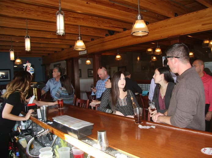 A group of people standing and chatting at a wooden bar with hanging lights. A female bartender serves drinks. The atmosphere is relaxed and social.