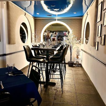 A narrow restaurant dining area with a high table and bar stools. Circular mirrors and glowing lights adorn the walls. Decorative white plants are placed at the end of the aisle. The ceiling is blue, and the floor is tiled.