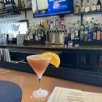 A cocktail in a martini glass with an orange slice garnish sits on a bar counter. Behind it are various liquor bottles and a television mounted on the wall above. A menu is partially visible on the counter.
