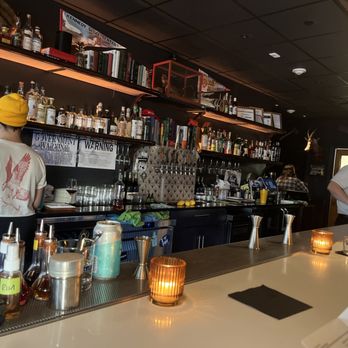 A dimly lit bar with bottles on shelves, glasses hanging overhead, and a bartender in a yellow hat. The counter has candles and drink-making tools. The atmosphere is cozy and intimate.