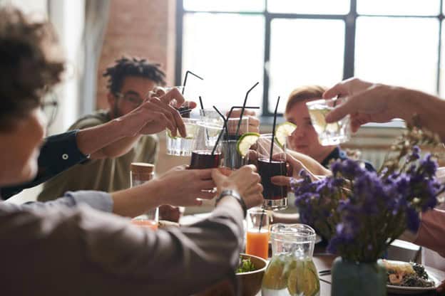 A group of people seated around a table raise glasses in a toast. The table is set with assorted drinks, a vase of purple flowers, and a bowl of greens. Sunlight filters through a large window in the background.