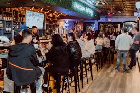 A lively bar scene with people sitting on stools and standing around a long bar counter. A bartender is serving drinks, and a TV screen is mounted above, displaying a sports game. The interior is warmly lit with wooden flooring and shelves.