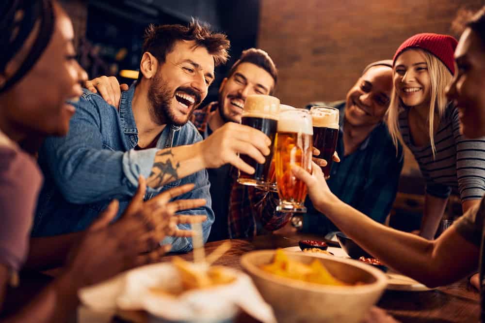 A group of friends cheerfully clinking glasses of beer at a bar. They are sitting around a wooden table with snacks, smiling and laughing. The atmosphere is warm and lively.
