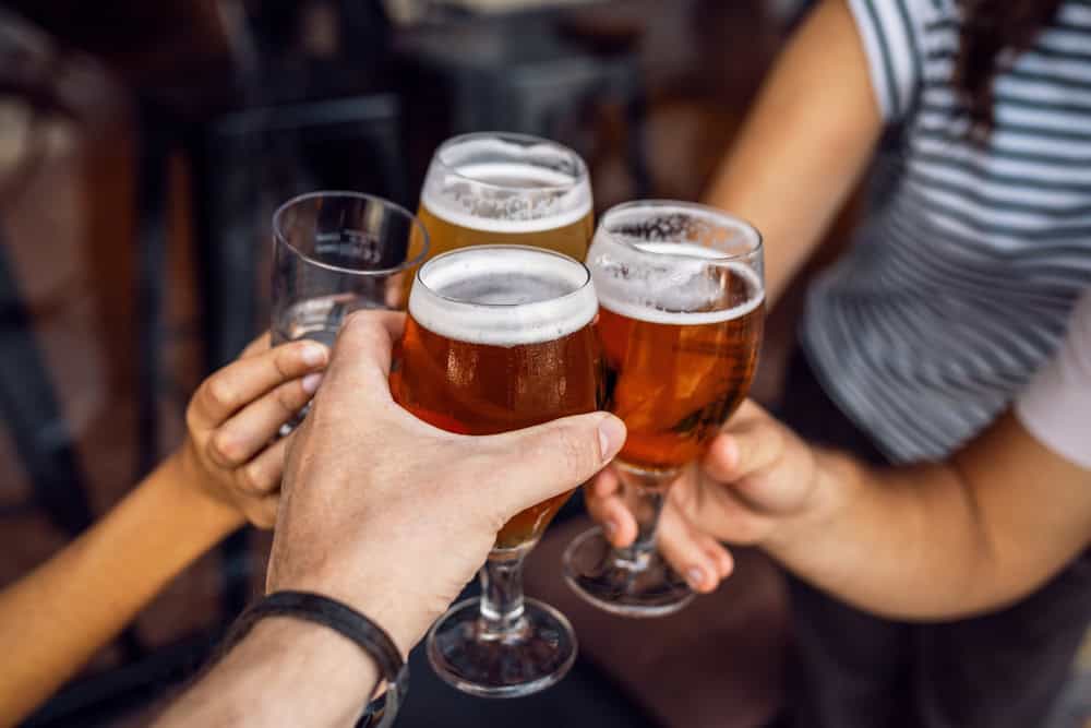 Four people clink their beer glasses together in a toast. Three glasses contain amber-colored beer, and one has a darker brew. The setting appears casual, with one person wearing a striped shirt and another with a wristband.