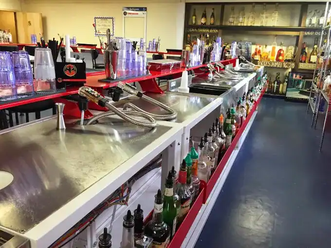 A modern bar setup with metal countertops, various bottles of liquor organized below, drink dispensers, stacked plastic cups, and shelves filled with more spirits in the background. The space appears clean and ready for service.