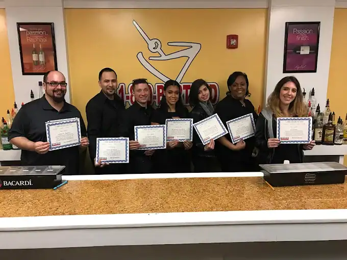 Seven people stand behind a bar holding certificates and smiling. Bottles and bar equipment are visible behind them, and a yellow wall has a logo with a martini glass and the word “HAR”.