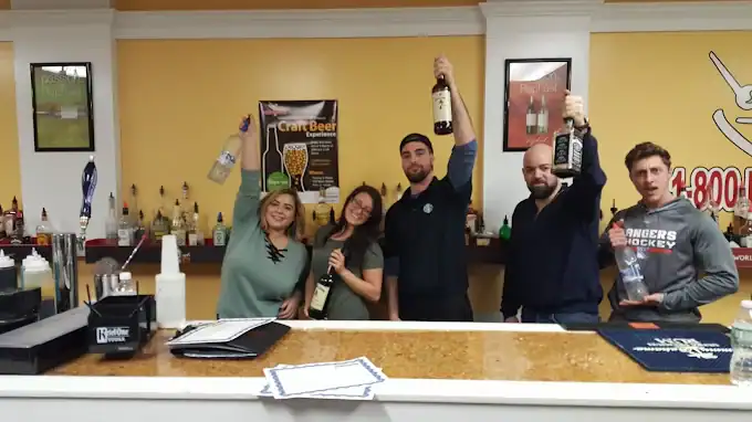 Five people stand behind a bar, smiling and holding up bottles of liquor. The bar counter has papers and bar supplies, and various bottles and posters are visible on the wall behind them.