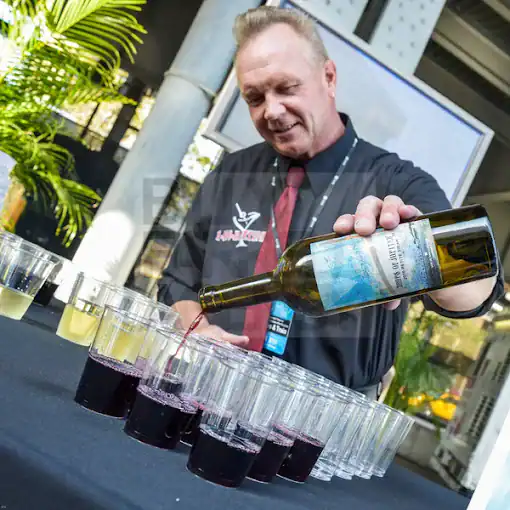 A man in a black shirt and red tie pours red wine from a bottle into plastic cups arranged on a table, with a few cups of white wine beside them. There are plants and pillars in the background.