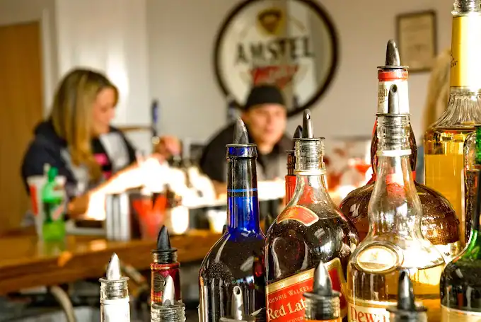 Several liquor bottles with pour spouts are in focus on a bar counter, while two people sit and talk in the blurred background. A large Amstel sign hangs on the wall behind them.
