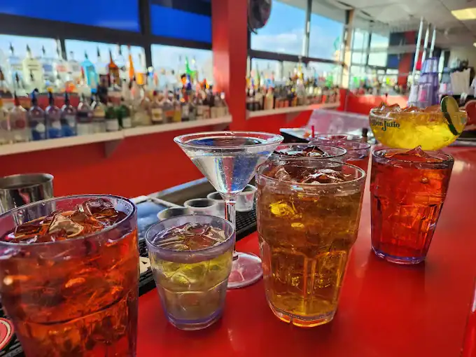 Five colorful cocktails and a martini glass are arranged on a red bar counter, with a variety of liquor bottles lined up on shelves in the background and sunlight streaming through large windows.