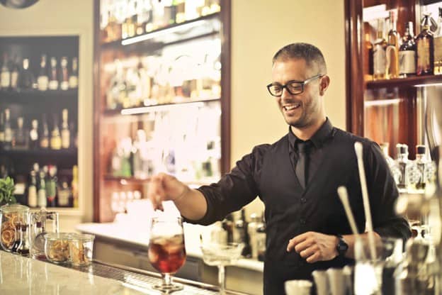 A smiling bartender in a black vest, shirt, and tie stirs a drink at a well-stocked bar, surrounded by bottles and bar tools, with warm lighting in the background.