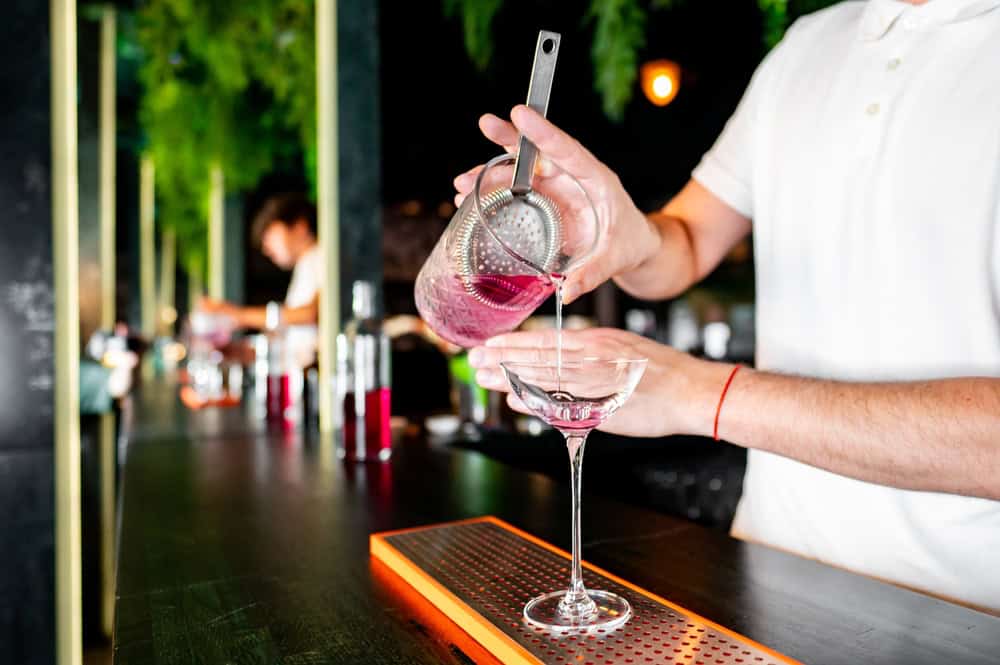 A bartender in a white shirt pours a pink cocktail from a mixing glass through a strainer into a coupe glass at a modern bar with bottles visible in the background.