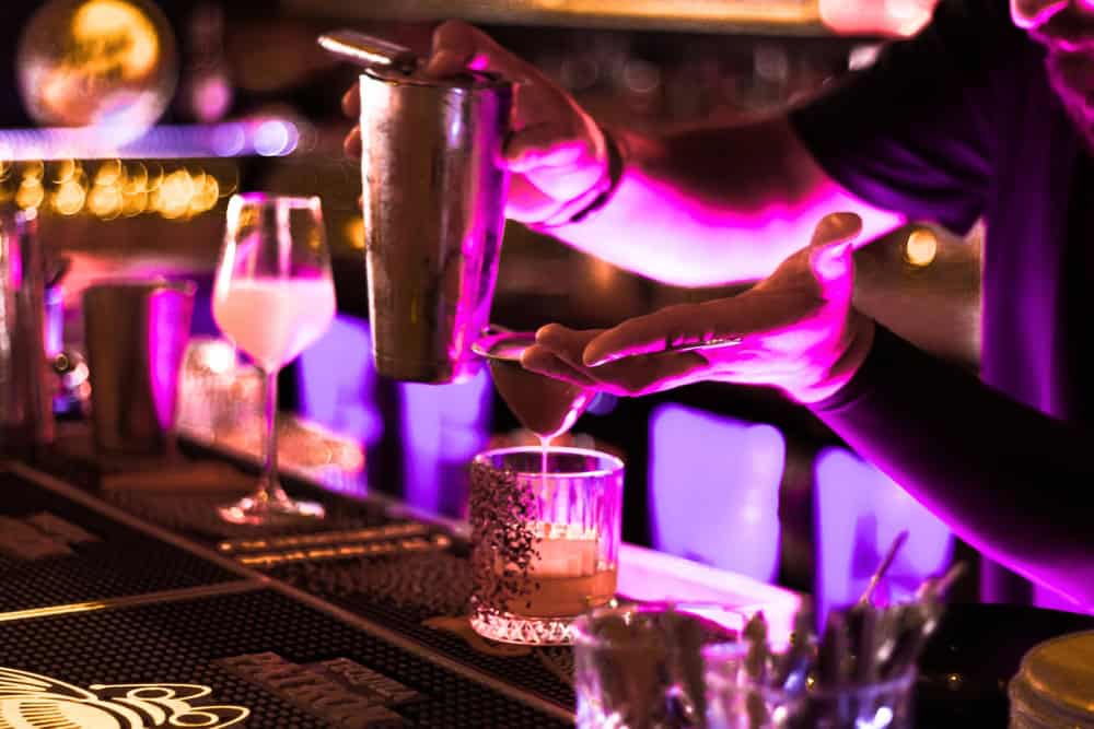 Bartender pours a drink from a shaker into a glass at a bar with vibrant purple and yellow lighting, surrounded by cocktail glasses and bar tools.