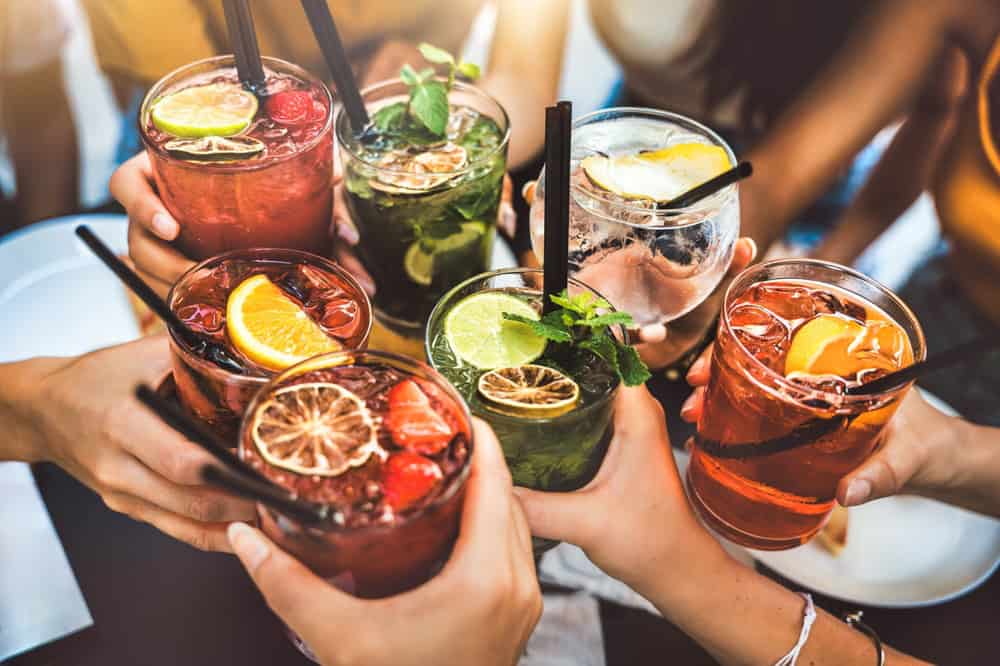 A group of people clinking colorful cocktails and mocktails garnished with fruit slices, berries, and mint leaves, creating a festive and cheerful atmosphere.