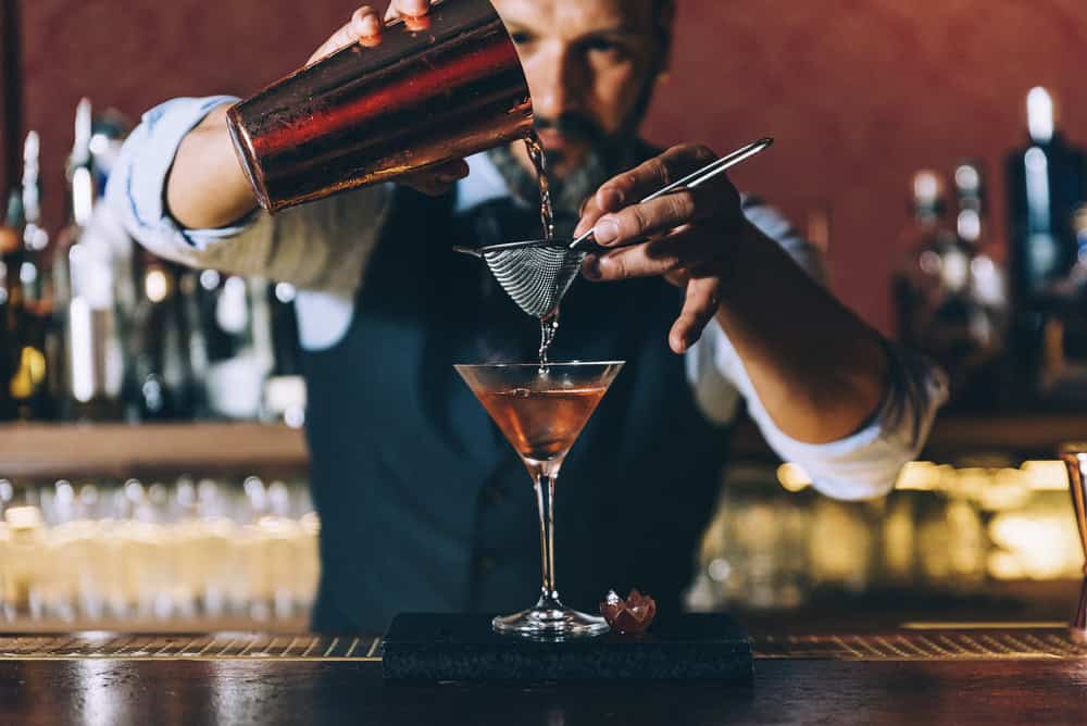 A bartender pours a cocktail through a strainer into a martini glass at a dimly lit bar, with bottles visible in the blurred background.