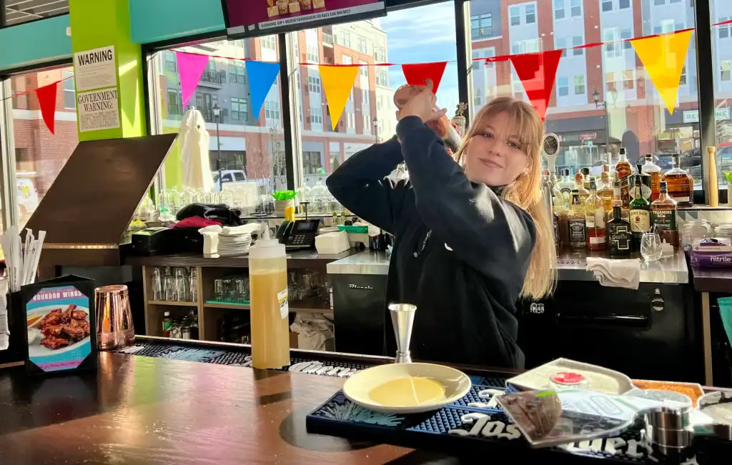 A bar counter with various bottles of liquor and mixers arranged on shelves in the background. The counter has a drink preparation area with a jigger, a plate, a bottle of mixer, and colorful triangular flags hanging above the window, which shows a view of buildings outside.