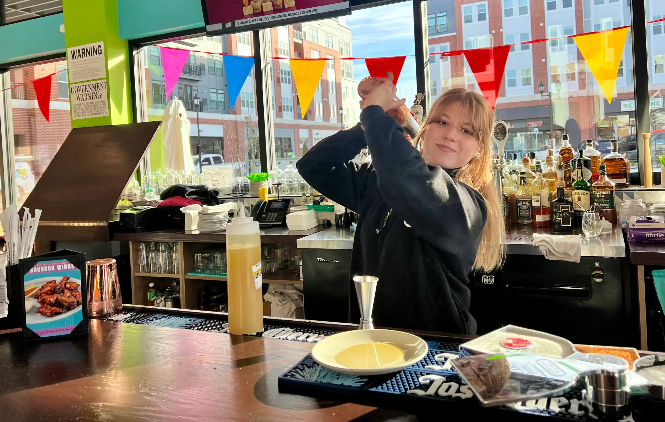 A bar counter with various bottles of liquor and mixers arranged on shelves in the background. The counter has a drink preparation area with a jigger, a plate, a bottle of mixer, and colorful triangular flags hanging above the window, which shows a view of buildings outside.